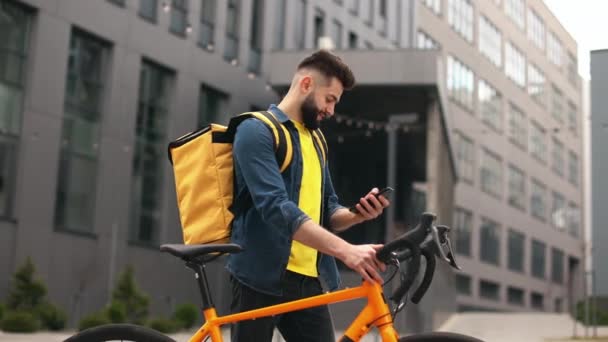 Ten poslíček s jídlem stojí a usmívá se. Dívá se na smartphone. Drží kolo a na ramenou má velký batoh. Kamera se pohybuje. Krásná práce — Stock video