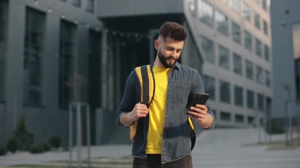 Der Essenslieferant steht und schaut auf das Smartphone. Er trägt einen großen Rucksack auf den Schultern. Er schreibt SMS. Schöne Business Center im Hintergrund. 4K — Stockvideo