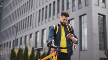 Yemek dağıtan adam ayakta duruyor ve akıllı telefona bakıyor. O bir bisiklet tutuyor ve omuzlarında büyük bir sırt çantası taşıyor. Kamera etrafta dolanıyor. Portre çekimi. 4K