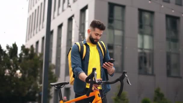 Ten poslíček stojí a píše smsky. Dívá se na smartphone. Drží kolo a na ramenou má velký batoh. Kamera se pohybuje. Portrét natáčení — Stock video