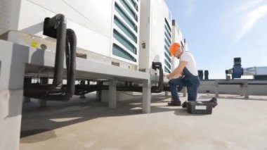 Genç mühendis havalandırma sistemini koruyor. Anahtarla vidayı çeviriyor. İş merkezinin çatısında yer almaktadır. Onun yanında bir alet çantası var. İş elbisesi giyiyor.