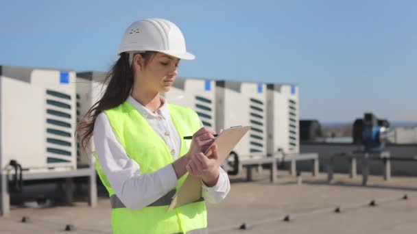 Portret skoncentrowanej inżynierki, która dokonuje wpisów w dokumentach i przegląda system klimatyzacji. Jest na dachu centrum biznesowego. Ma na sobie ubranie robocze i twardy kapelusz. — Wideo stockowe