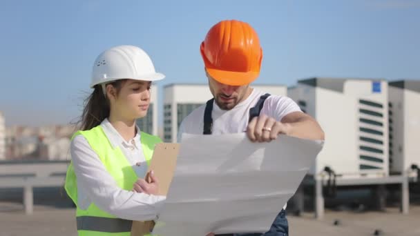 Dwóch inżynierów omawia rysunki projektów. Są na dachu centrum biznesowego. Trzymają w rękach dokumenty i rysunki. Noszą ubrania robocze i twardy kapelusz. Powietrze — Wideo stockowe