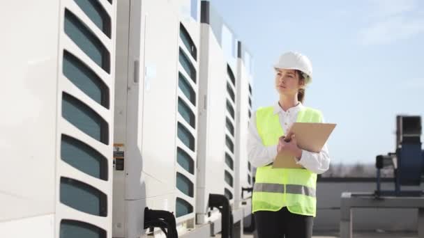 Μια νεαρή γυναίκα μηχανικός εξετάζει τα κλιματιστικά. Κρατάει σημειώσεις στα έγγραφα. Είναι στην οροφή ενός εμπορικού κέντρου. Φοράει ρούχα εργασίας και ένα σκληρό καπέλο. Ωραία ηλιόλουστη μέρα. 4K — Αρχείο Βίντεο