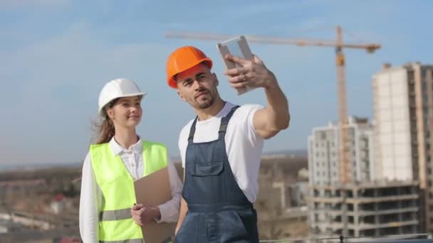 Два счастливых инженера улыбаются и делают селфи. Мужчина держит планшет в руке и фотографирует. Они на крыше бизнес-центра. На них рабочая одежда и каска. — стоковое видео
