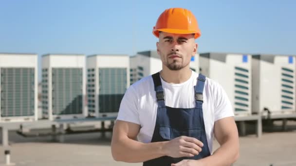 Porträtt av en seriös ung ingenjör som står och tittar på kameran. Han korsar armarna. Han är på taket till ett affärscenter. Han har arbetskläder och hatt. Luft — Stockvideo