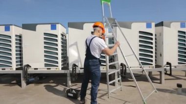 Genç mühendis bir merdiven diziyor ve onu tırmanıyor. Klimayı o çalıştırıyor. Bir iş merkezinin çatısında. Onun yanında bir alet çantası var. İş elbisesi giyiyor.