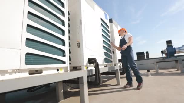 Een jonge ingenieur voert een inspectie uit en controleert de airconditioners met zijn handen. Hij draagt werkkleding en een harde hoed. Airconditioning op de achtergrond. Hij zit op het dak. — Stockvideo