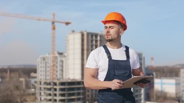 若いエンジニアは書類とスマートフォンのフォルダを保持しています。彼はカメラを真剣に見ている。彼は仕事着と堅い帽子をかぶっている。彼はビジネスセンターの屋上にいる — ストック動画