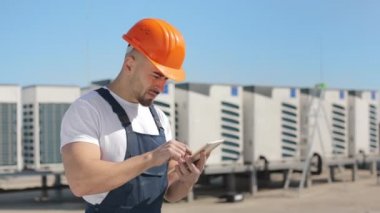 Ciddi bir genç mühendis tablet üzerinde çalışıyor ve kameraya bakıyor. O, iş kıyafetleri ve sert bir şapka giyiyor. Arka planda havalandırma sistemleri var. Güzel güneşli bir gün. 4K