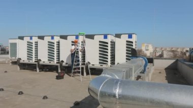 Genç mühendis merdivenleri tırmanıyor ve vidaları anahtarla çeviriyor. Bir iş merkezinin çatısında. O, iş kıyafetleri ve sert bir şapka giyiyor. Güzel güneşli bir gün. 4K