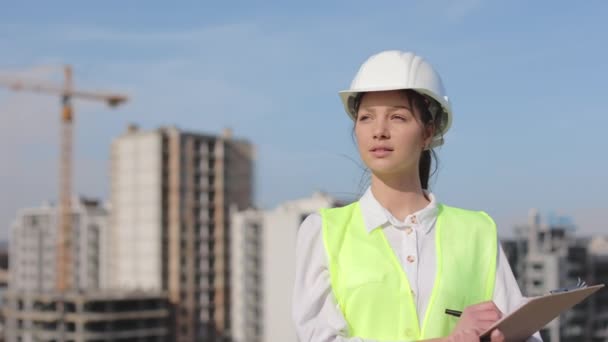 Kameraya bakan bir kadın mühendisin portresi. Saçları rüzgarda uçuşuyor. O, iş kıyafetleri ve sert bir şapka giyiyor. Bir iş merkezinin çatısında. İnşaat... — Stok video
