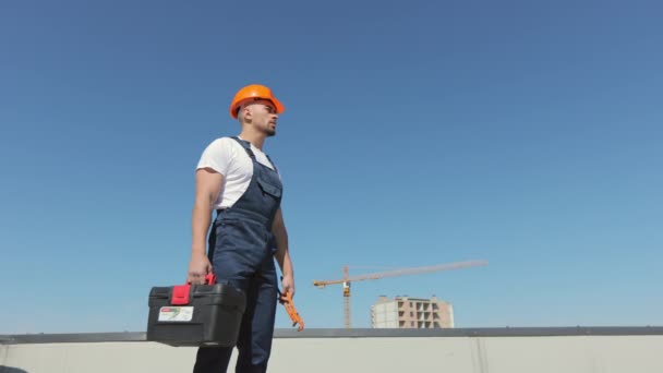 Un ingeniero serio sostiene una caja de herramientas y una llave de tubo en su mano. Está examinando el techo. Lleva ropa de trabajo y un sombrero duro. Construcción en el fondo. Bonito día soleado. 4K — Vídeos de Stock