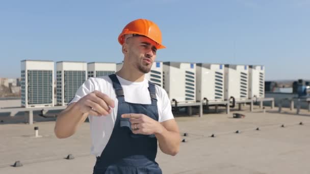 De gelukkige ingenieur kijkt naar de camera en praat emotioneel. Hij zwaait met zijn armen en praat over het airconditioningsysteem. Hij draagt werkkleding. Hij draagt werkkleding en een — Stockvideo