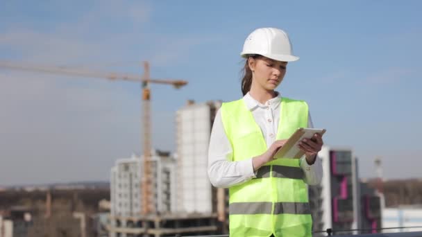 Portrait de jeune femme ingénieur qui travaille sur la tablette. Ses cheveux sont époustouflés par le vent. Elle est sur le toit d'un centre d'affaires. Elle porte des vêtements de travail et un casque. Construction — Video