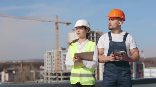 Zwei seriöse Ingenieure stehen und schauen weg. Sie tragen Arbeitskleidung und einen harten Hut. Sie befinden sich auf dem Dach eines Geschäftszentrums. Bau im Hintergrund. Schöner sonniger Tag. 4K — Stockvideo