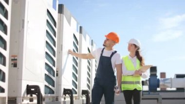 İki mutlu mühendis klimaların yanından geçiyor ve teftişlerini yapıyorlar. Gülümsüyorlar. Bir iş merkezinin çatısındalar. İş kıyafetleri ve kalın bir şapka giyiyorlar. Güzel.
