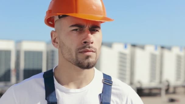 Portret van een serieuze jonge ingenieur die zijn hoofd draait en naar de camera kijkt. Hij zit op het dak van een zakencentrum. Hij draagt werkkleding en een harde hoed. Luchtbehandelingssystemen in — Stockvideo