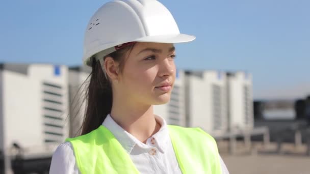 Portret szczęśliwej inżynierki patrzy w kamerę. Ma na sobie ubranie robocze i twardy kapelusz. Jest na dachu centrum biznesowego. Klimatyzacja w tle. Ładne słońce. — Wideo stockowe