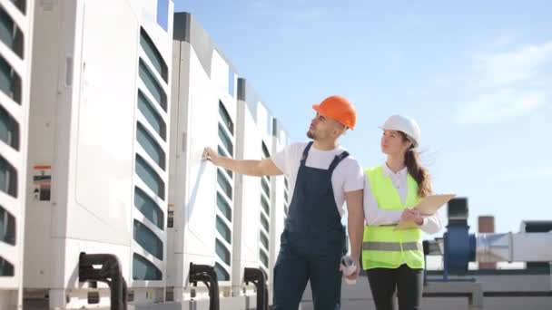 Dwóch szczęśliwych inżynierów przechodzi obok klimatyzatorów i przeprowadza inspekcję. Uśmiechają się. Są na dachu centrum biznesowego. Noszą ubrania robocze i twardy kapelusz. Ładnie. — Wideo stockowe