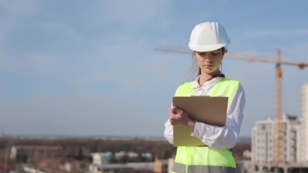 Portrait d'une jeune ingénieure qui fait des entrées dans des documents. Elle est sur le toit d'un centre d'affaires. Elle porte des vêtements de travail et un casque. Construction en arrière-plan. Belle journée ensoleillée — Video