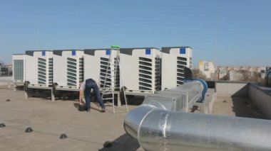 Genç bir mühendis alet kutusunu açıp boru anahtarını alıyor. Bir iş merkezinin çatısında. Onun yanında bir merdiven duruyor. O, iş kıyafetleri ve sert bir şapka giyiyor. Güzel güneşli bir gün. 4K