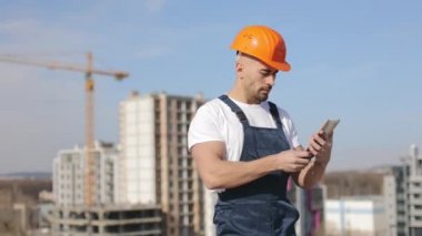 Tablet üzerinde çalışan genç bir mühendisin portresi. Başını çeviriyor ve kameraya bakıyor. Bir iş merkezinin çatısında. İş kıyafetleri ve sıkı bir şapka giyiyor.