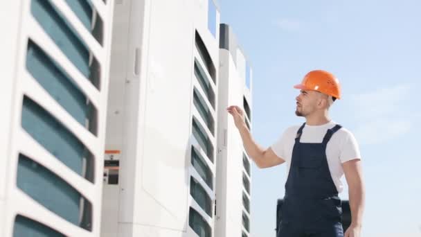 Een jonge ingenieur gaat het airconditioningsysteem controleren. Hij onderhoudt het airco systeem. Hij zit op het dak van een zakencentrum. Hij draagt werkkleding en een harde hoed. Mooi. — Stockvideo