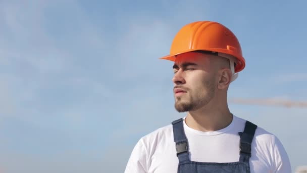 Portret van een gelukkige ingenieur die naar de camera kijkt. Hij draagt werkkleding en een harde hoed. Hij is op het dak van een zakencentrum. Constructie op de achtergrond. Mooie zonnige dag. 4K — Stockvideo