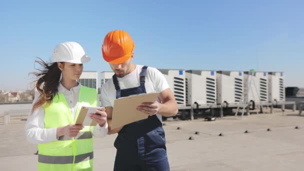 Dois engenheiros felizes estão discutindo documentos de trabalho e fazendo anotações. Um vento forte está soprando. Eles estão usando roupas de trabalho e um chapéu duro. Eles estão no telhado de um centro de negócios. Ar condicionado — Vídeo de Stock