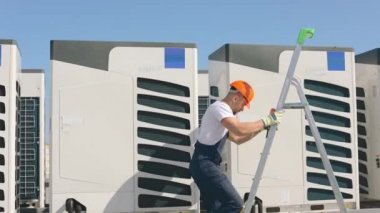 Genç mühendis merdivenleri tırmanıyor ve bir boru anahtarıyla vidaları çeviriyor. Klimayı o çalıştırıyor. O, iş kıyafetleri ve sert bir şapka giyiyor. Güzel güneşli bir gün. 4K