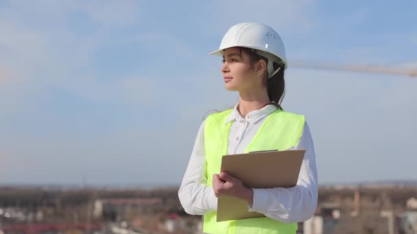 Başka tarafa bakan bir kadın mühendisin portresi. Saçları rüzgarda uçuşuyor. O, iş kıyafetleri ve sert bir şapka giyiyor. Bir iş merkezinin çatısında. Arkaplanda inşaat — Stok video