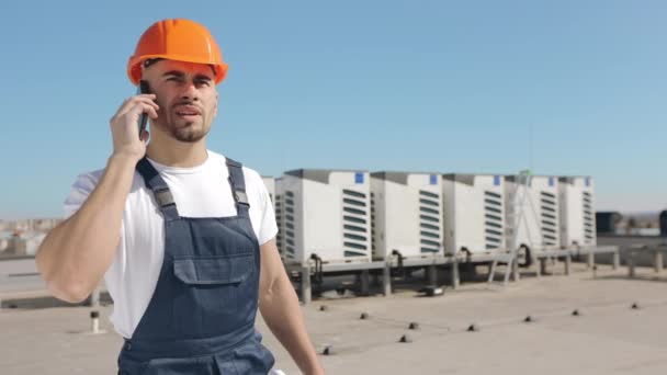 Retrato de un joven ingeniero serio que está discutiendo temas de trabajo por teléfono y cuelga. Él sostiene un dibujo del proyecto en sus manos. Lleva ropa de trabajo y un sombrero duro. Aire acondicionado — Vídeos de Stock