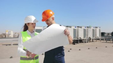 İki mutlu mühendis proje çizimlerini tartışıyor. Gülümsüyorlar. Bir iş merkezinin çatısındalar. İş kıyafetleri ve kalın bir şapka giyiyorlar. Havalandırma sistemi