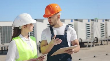 İki mutlu mühendis çalışma belgelerini tartışıyor ve notlar alıyorlar. Rüzgar esiyor. İş kıyafetleri ve kalın bir şapka giyiyorlar. Arka planda havalandırma sistemi var. 4K