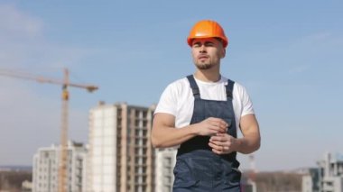 Genç mühendis kameraya bakıyor ve kollarını kavuşturuyor. O, iş kıyafetleri ve sert bir şapka giyiyor. Arka planda inşaat var. Güzel güneşli bir gün. 4K