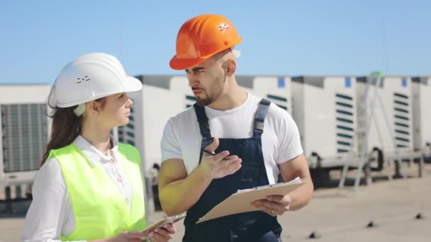 Dos ingenieros felices están discutiendo documentos de obras y tomando notas. Un viento sopla. Llevan ropa de trabajo y un sombrero duro. Sistema de aire acondicionado en el fondo. 4K — Vídeo de stock