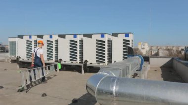 Genç mühendis bir merdiven ve alet çantasıyla klimalar için uygundur. O, merdiveni açıyor. Bir iş merkezinin çatısında. O, iş kıyafetleri ve sert bir şapka giyiyor. Güzel güneşli