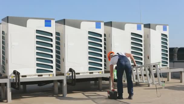 Genç mühendis bir merdiven ve alet çantasıyla klimalar için uygundur. Anahtarı alıyor ve merdivenleri çıkıyor. Klimayı o çalıştırıyor. O bir çatıda. — Stok video