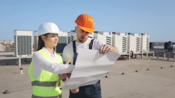 Een jonge vrouwelijke ingenieur legt de projectgegevens uit aan de ingenieur. Ze zijn op het dak van een zakencentrum. Ze dragen werkkleding en een harde hoed. Airconditioningsystemen op de achtergrond — Stockvideo