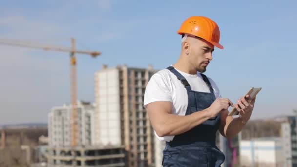 Portrét mladého inženýra, který pracuje na desce. Je na střeše obchodního centra. Má na sobě pracovní oblečení a tvrdý klobouk. Stavba v pozadí. 4K — Stock video