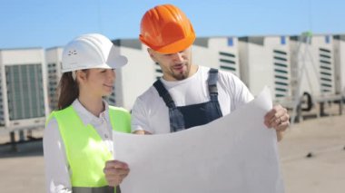 Two happy engineers are discussing project drawings. They are smiling. They are wearing work clothes and a hard hat. Air conditioning system in the background. Nice sunny day. 4K