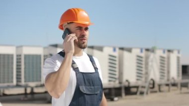 A young serious engineer is talking on the phone. He is wearing work clothes and a hard hat. Air conditioning system in the background. 4K