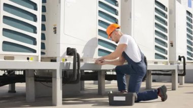 The young concentrated engineer is maintaining the air conditioning system. He is turning the screw with a pipe wrench. Hes wearing work clothes and a hard hat. He is located on the roof of the