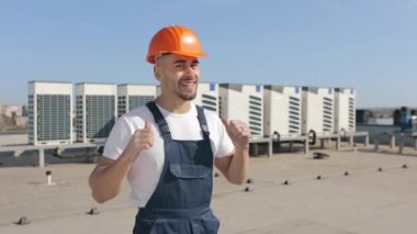 The happy engineer is looking at the camera and showing two fingers up. He is wearing work clothes. He is wearing work clothes and a hard hat. Air conditioning systems in the background. Nice sunny
