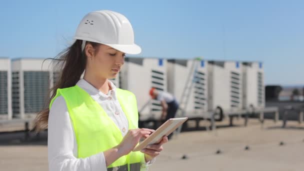 Portrait of a young woman engineer who is working on a tablet and watching the engineers work. The engineer is climbing a ladder in the background. They are wearing work clothes and a hard hat. Air — Video