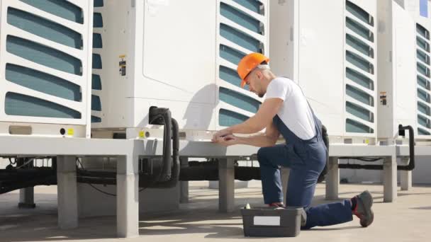 The young concentrated engineer is maintaining the air conditioning system. He is turning the screw with a pipe wrench. Hes wearing work clothes and a hard hat. He is located on the roof of the — ストック動画