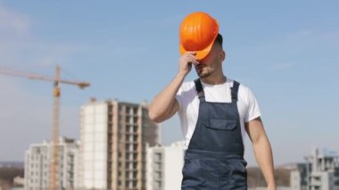 The young engineer is putting on a protective helmet on his head and looking at the camera. He is putting his hands on his belt. He is wearing work clothes. Construction in the background. Nice sunny