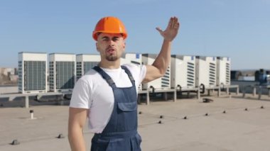 The engineer is emotionally talking about the air conditioning system and looking into the camera. He smiling and showing a thumb up. Hes on the roof of a business center. He is wearing work clothes