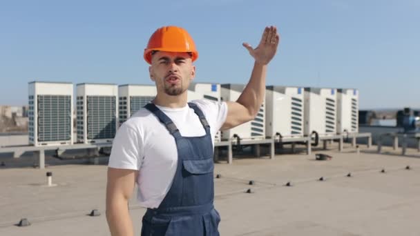 Ingenjören pratar känslomässigt om luftkonditioneringssystemet och tittar in i kameran. Han ler och visar tummen upp. Han är på taket till ett affärscenter. Han har arbetskläder. — Stockvideo